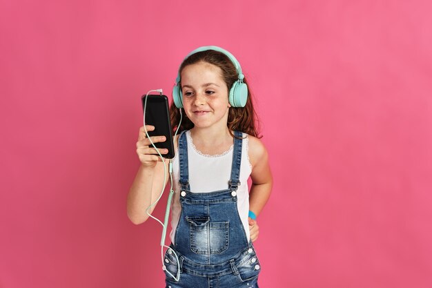 Niña linda posando con un teléfono y auriculares en una pared rosa