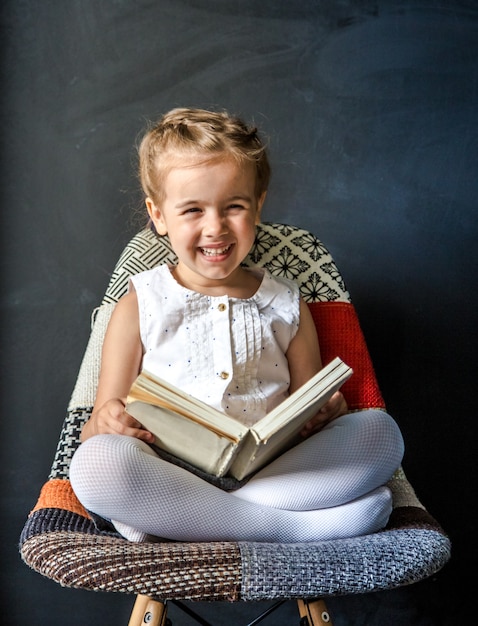 Niña linda sentada en una silla hermosa con un libro en la mano, el concepto de educación y vida escolar