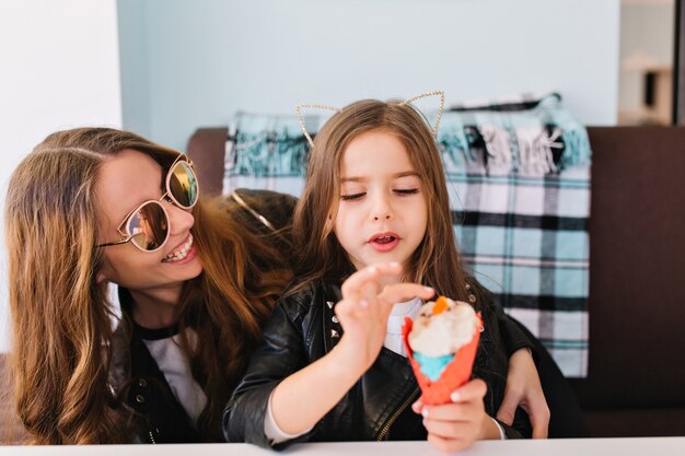 Niña linda y su atractiva mamá alegre con gafas de sol de moda divirtiéndose en casa y comiendo postre.