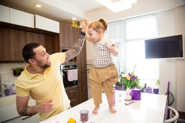 Niña linda con su padre feliz y alegre sosteniendo su brazo sobre la mesa de la cocina tratando de dar un paso divirtiéndose juntos en la cocina en casa concepto de familia feliz