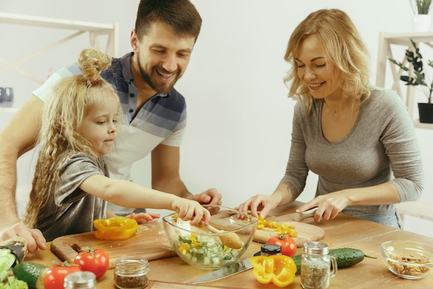 Niña linda y sus hermosos padres están cortando verduras en la cocina de casa