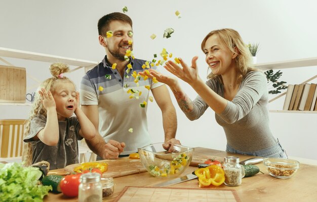 Niña linda y sus hermosos padres están cortando verduras en la cocina de casa