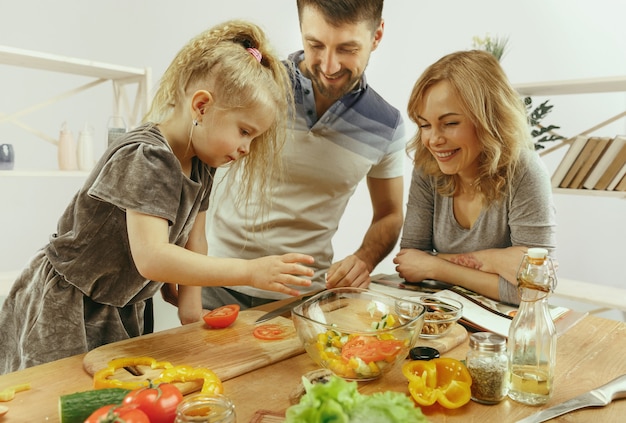 Niña linda y sus hermosos padres están cortando verduras en la cocina de casa