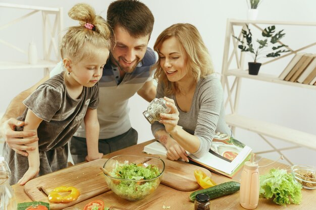 Niña linda y sus hermosos padres están cortando verduras en la cocina de casa