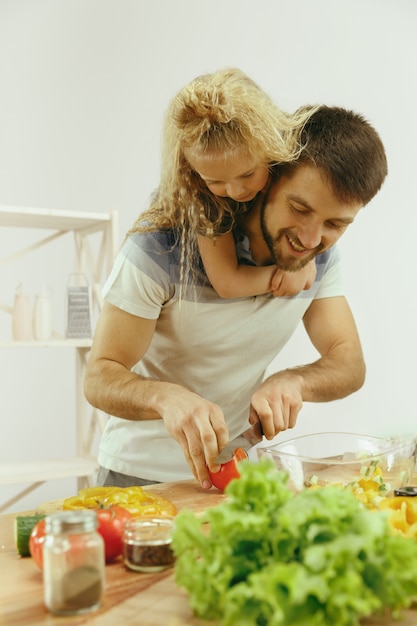 Niña linda y sus hermosos padres están cortando verduras y sonriendo mientras hacen ensalada en la cocina de casa. Concepto de estilo de vida familiar