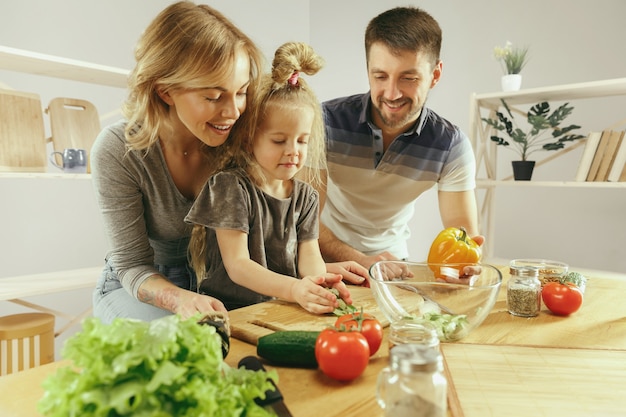 Niña linda y sus hermosos padres están cortando verduras y sonriendo mientras hacen ensalada en la cocina de casa. Concepto de estilo de vida familiar