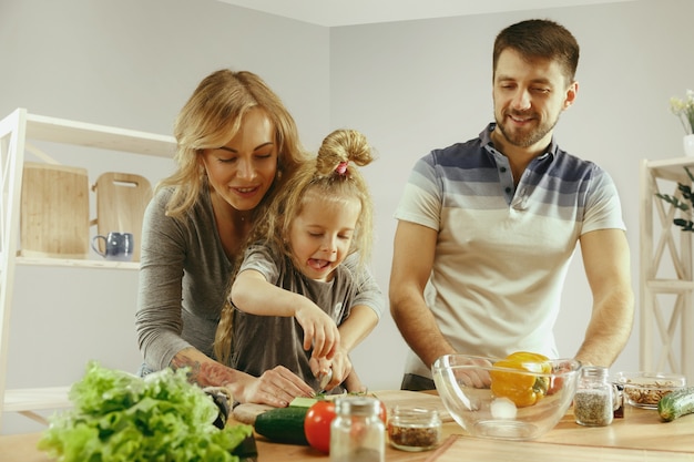 Niña linda y sus hermosos padres están cortando verduras y sonriendo mientras hacen ensalada en la cocina de casa. Concepto de estilo de vida familiar