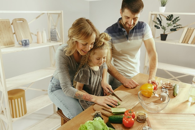 Niña linda y sus hermosos padres están cortando verduras y sonriendo mientras hacen ensalada en la cocina de casa