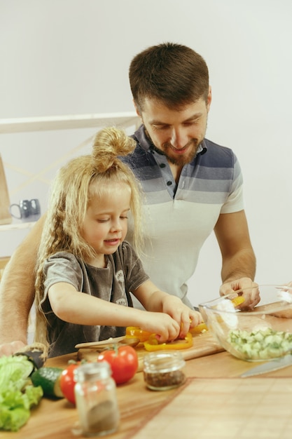 Niña linda y sus hermosos padres están cortando verduras y sonriendo mientras hacen ensalada en la cocina de casa