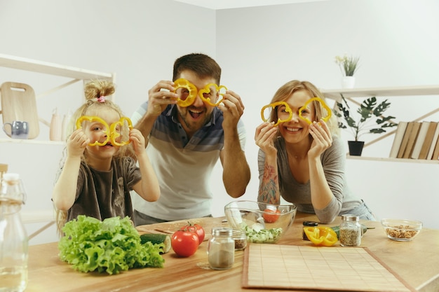 Niña linda y sus hermosos padres están cortando verduras y sonriendo mientras hacen ensalada en la cocina de casa