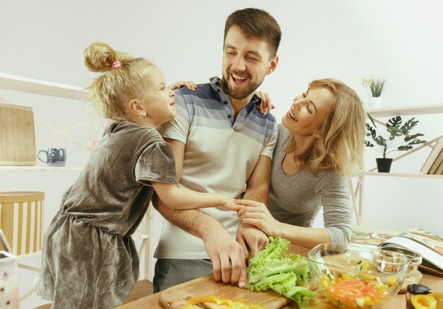 Niña linda y sus hermosos padres están cortando verduras y sonriendo mientras hacen ensalada en la cocina de casa