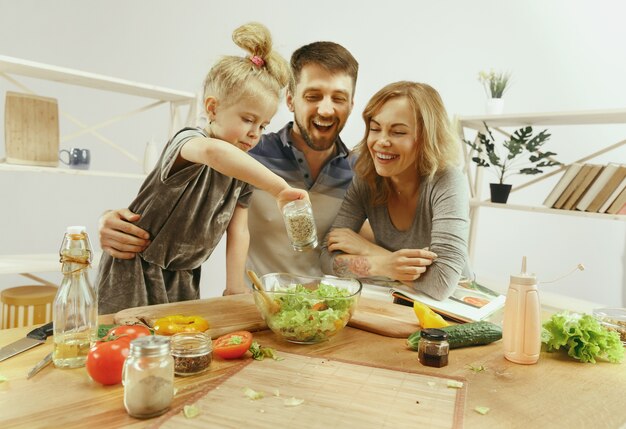 Niña linda y sus hermosos padres están cortando verduras y sonriendo mientras hacen ensalada en la cocina de casa