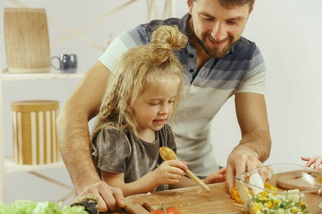 Niña linda y sus hermosos padres están cortando verduras y sonriendo mientras hacen ensalada en la cocina de casa