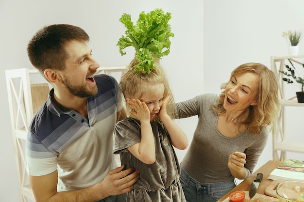 Niña linda y sus hermosos padres están cortando verduras y sonriendo mientras hacen ensalada en la cocina de casa