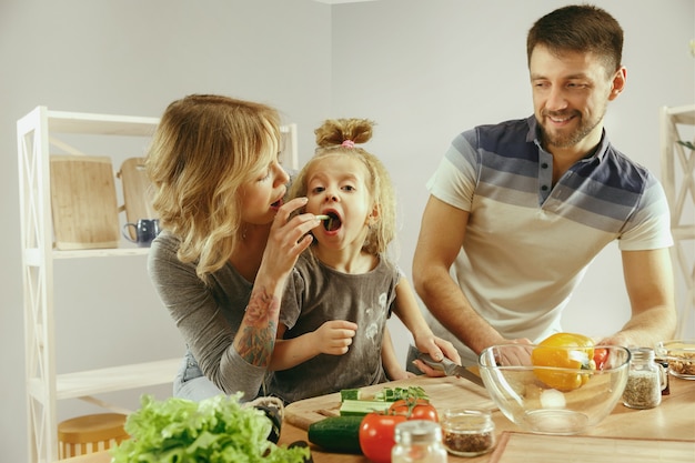 Niña linda y sus hermosos padres están cortando verduras y sonriendo mientras hacen ensalada