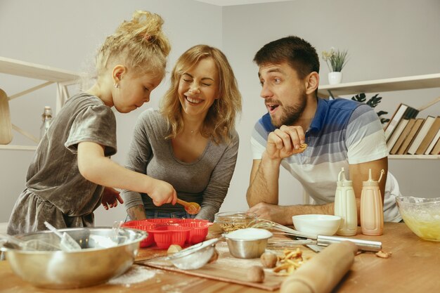 Niña linda y sus hermosos padres preparando la masa para el pastel en la cocina de casa. Concepto de estilo de vida familiar