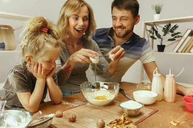 Niña linda y sus hermosos padres preparando la masa para el pastel en la cocina de casa. Concepto de estilo de vida familiar