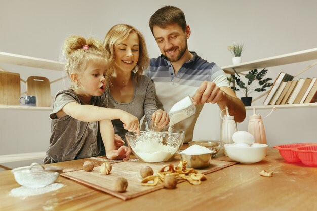 Niña linda y sus hermosos padres preparando la masa para el pastel en la cocina de casa