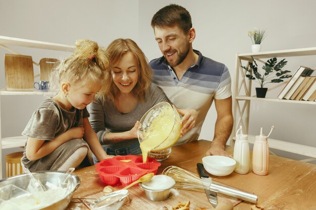 Niña linda y sus hermosos padres preparando la masa para el pastel en la cocina de casa.