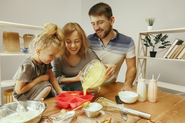 Niña linda y sus hermosos padres preparando la masa para el pastel en la cocina de casa.