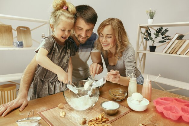 Niña linda y sus hermosos padres preparando la masa para el pastel en la cocina de casa