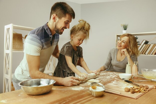 Niña linda y sus hermosos padres preparando la masa para el pastel en la cocina de casa.