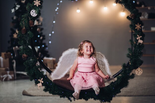 Una niña linda con un vestido con angel cruces se sienta en un gran escenario redondo de ramas de abeto