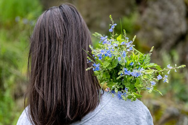 La niña lleva un ramo de flores recogidas en el bosque de primavera, vista desde atrás.