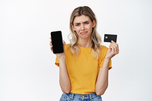 Niña llorando mostrando la pantalla vacía del teléfono móvil y la tarjeta de crédito, enfurruñada y decepcionada, no puede comprar algo, falta dinero en la cuenta bancaria, de pie en blanco