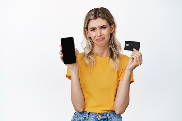 Niña llorando mostrando la pantalla vacía del teléfono móvil y la tarjeta de crédito, enfurruñada y decepcionada, no puede comprar algo, falta dinero en la cuenta bancaria, de pie en blanco