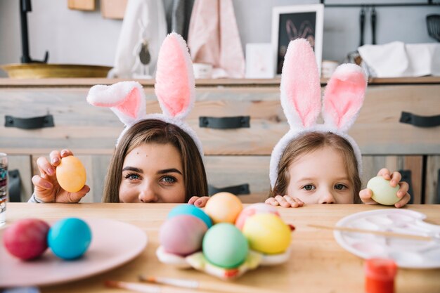 Niña y madre en orejas de conejo escondidas detrás de una mesa con huevos de colores