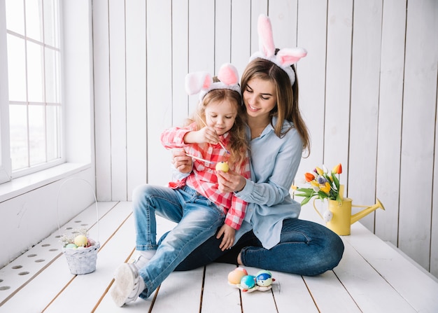 Niña y madre en orejas de conejo pintando huevos para Pascua