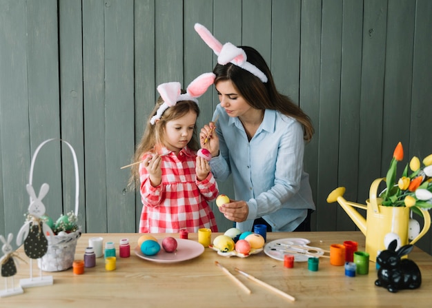 Foto gratuita niña y madre en orejas de conejo pintando huevos para pascua