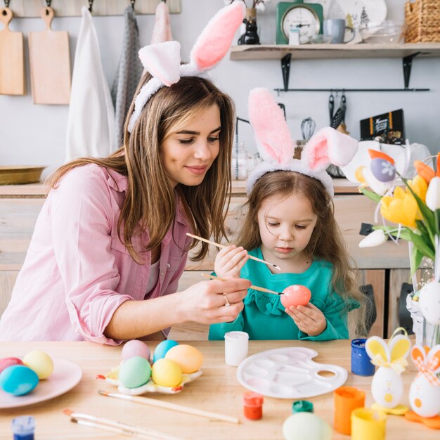 Niña con madre pintando huevos para Pascua en mesa