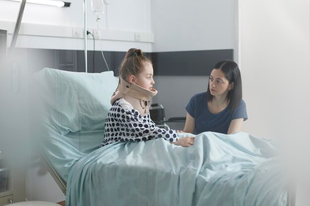 Niña malherida con collarín cervical hablando con una madre preocupada y ansiosa mientras se sienta en la cama del hospital. Hirió a la pequeña hija usando collar médico discutiendo con una mujer atenta y cariñosa.