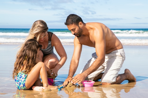 Niña con mamá y papá disfrutando de las vacaciones en el mar, jugando con juguetes en la arena mojada y en el agua