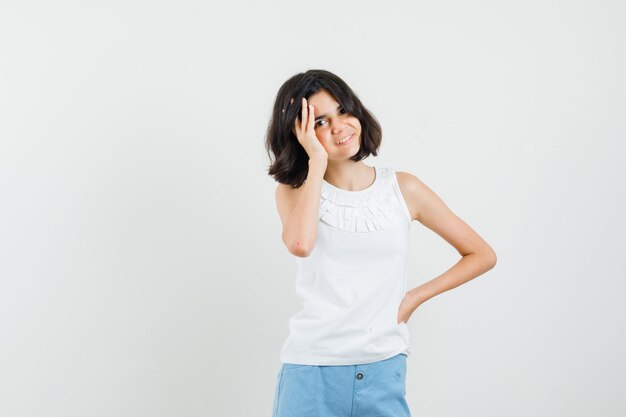 Niña de la mano a la cara en blusa blanca, pantalones cortos y mirando alegre, vista frontal.