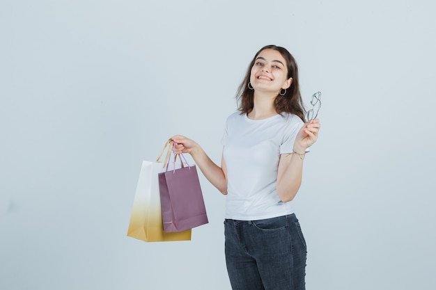 Niña manteniendo bolsas de regalo y vasos en camiseta, jeans y mirando feliz. vista frontal.