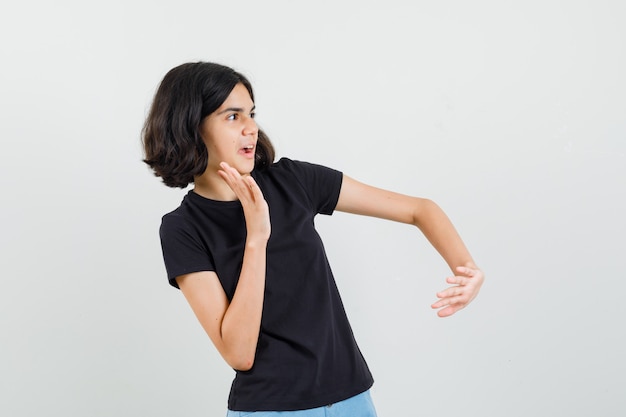 Niña manteniendo la mano de manera preventiva en camiseta negra, pantalones cortos y mirando asustada, vista frontal.