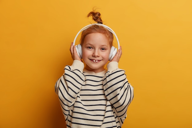 La niña meloman disfruta de un buen sonido con auriculares nuevos, escucha su canción favorita, se entretiene con música genial, muestra dientes de leche, usa un jersey de rayas, aislado en una pared amarilla, tiene un estado de ánimo despreocupado