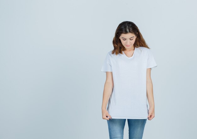 Niña mirando hacia abajo mientras sostiene la camiseta en la camiseta, jeans y parece confiada. vista frontal.