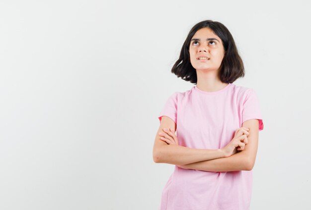 Niña mirando hacia arriba con los brazos cruzados en camiseta rosa y mirando pensativo, vista frontal.