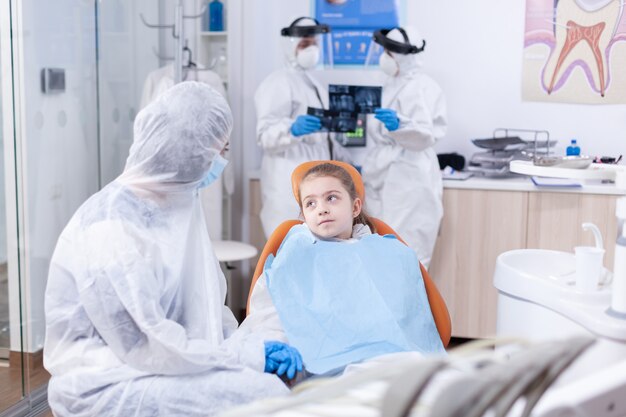 Niña mirando a una madre pensativa sentada en un sillón dental con un mono debido al brote de coronavirus. Estomatólogo durante covid19 vistiendo traje de ppe haciendo procedimiento de dientes de un niño sentado en una silla.