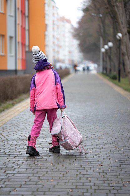 Niña con una mochila en una chaqueta y un sombrero cerca de la escuela.