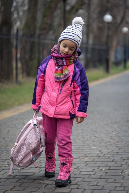 Niña con una mochila en una chaqueta y un sombrero cerca de la escuela.