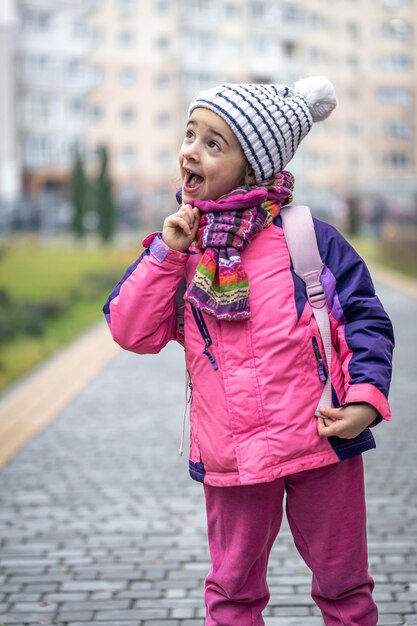 Niña con una mochila en una chaqueta y un sombrero cerca de la escuela.