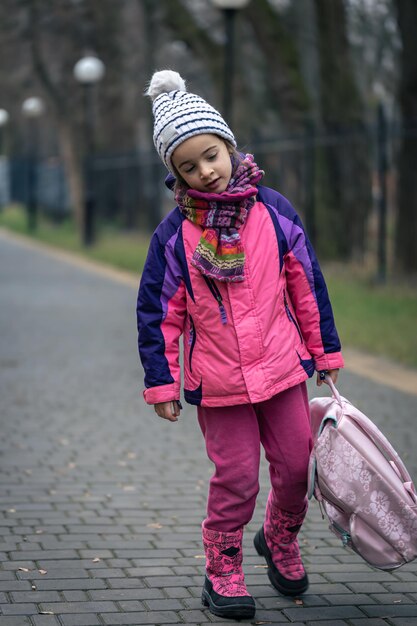 Niña con una mochila en una chaqueta y un sombrero cerca de la escuela.