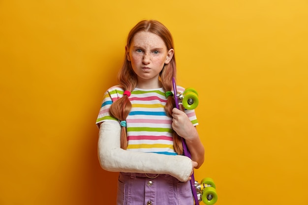 Niña molesta con cabello pelirrojo y pecas, cara sonriente y expresión insatisfecha, posa con patineta, no puede seguir conduciendo debido a un traumatismo en el brazo. Niños, salud, deporte de riesgo