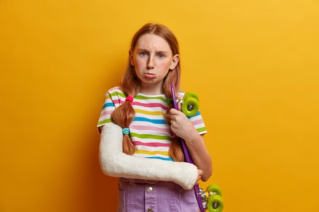 Niña molesta y ofendida que va a llorar, posa con patineta, quiere tener unas vacaciones de verano activas, se ha roto el brazo enyesado, lleva un estilo de vida deportivo, se ha roto el brazo después de hacer un truco peligroso