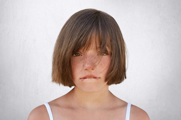 Niña molesta que disputa con sus padres, mirando inocentemente a la cámara mientras curva sus labios aislados en blanco. Sorprendente niña llorando, mordiéndose el labio inferior